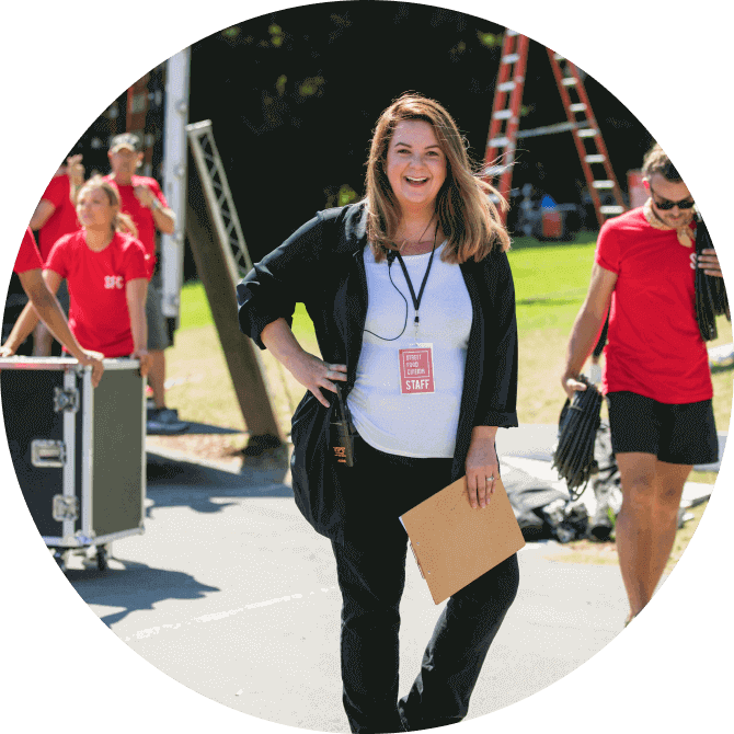 A non-profit manager poses for a photo at an outdoor event as her team works in the background.
